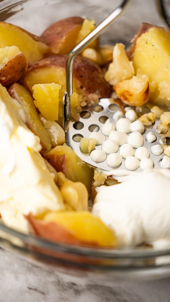 Red potatoes being mashed in a bowl with a potato masher, preparing creamy garlic mashed potatoes
